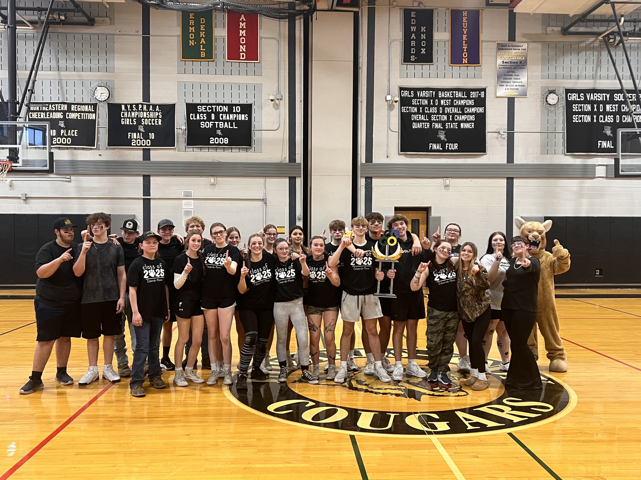 students in gym pointing one finger up, holding a trophy