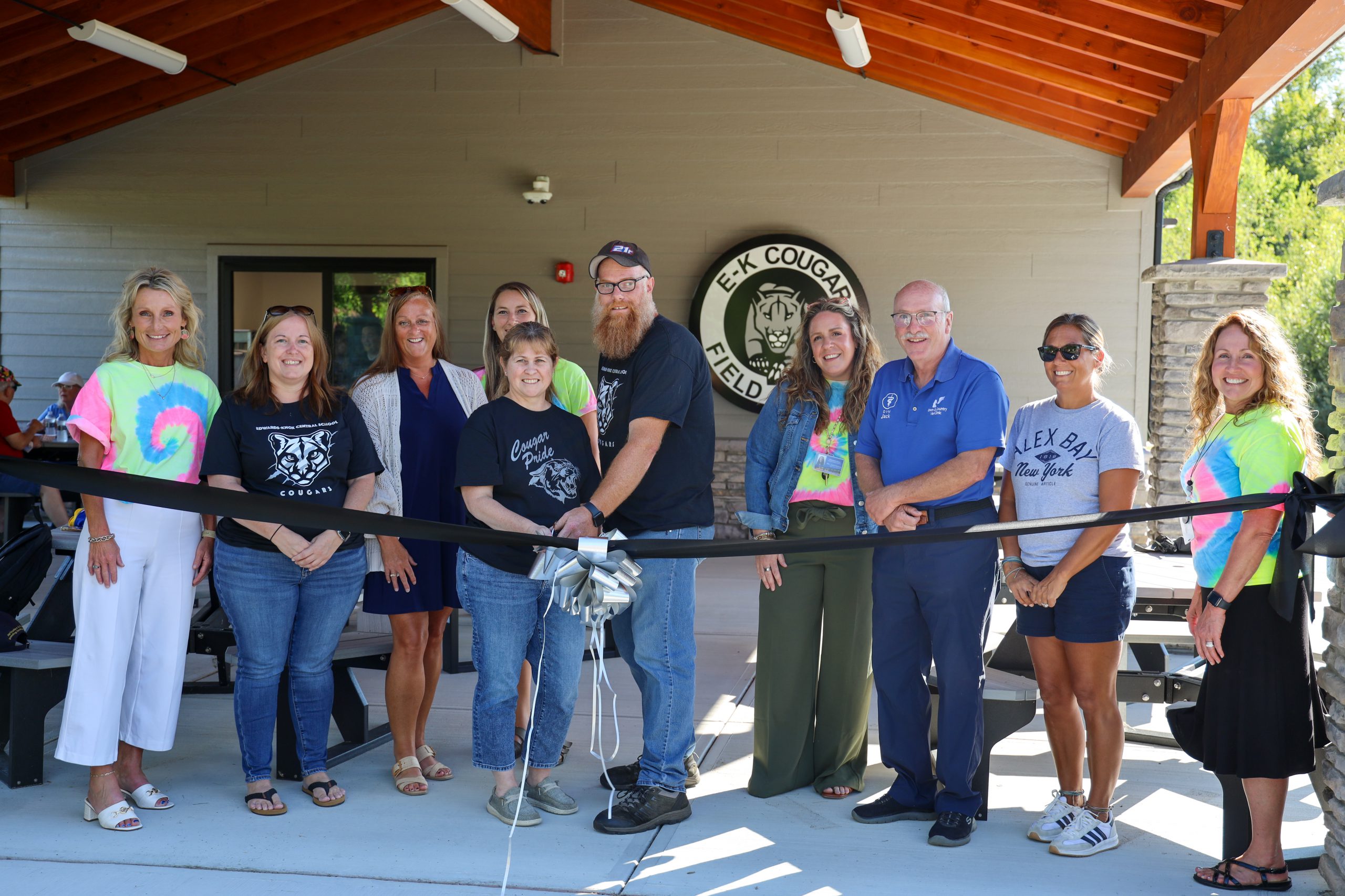 StudentTen people standing side by side with a ribbon and scissors in front of a field house